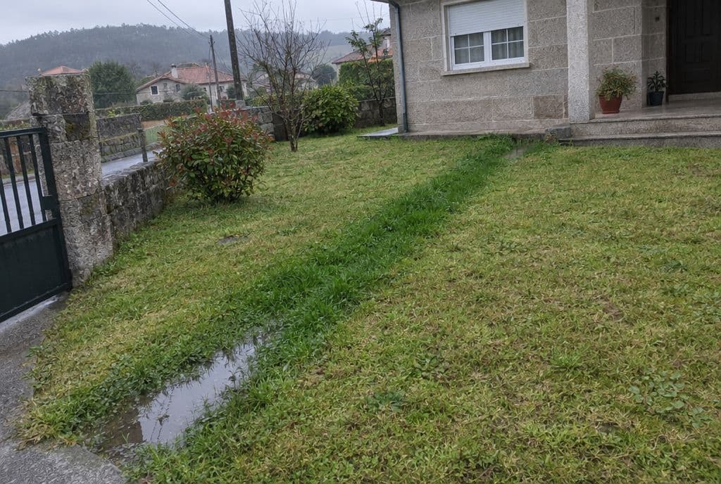 Franja de césped más verde y encharcada siguiendo el recorrido de una tubería enterrada en un jardín gallego