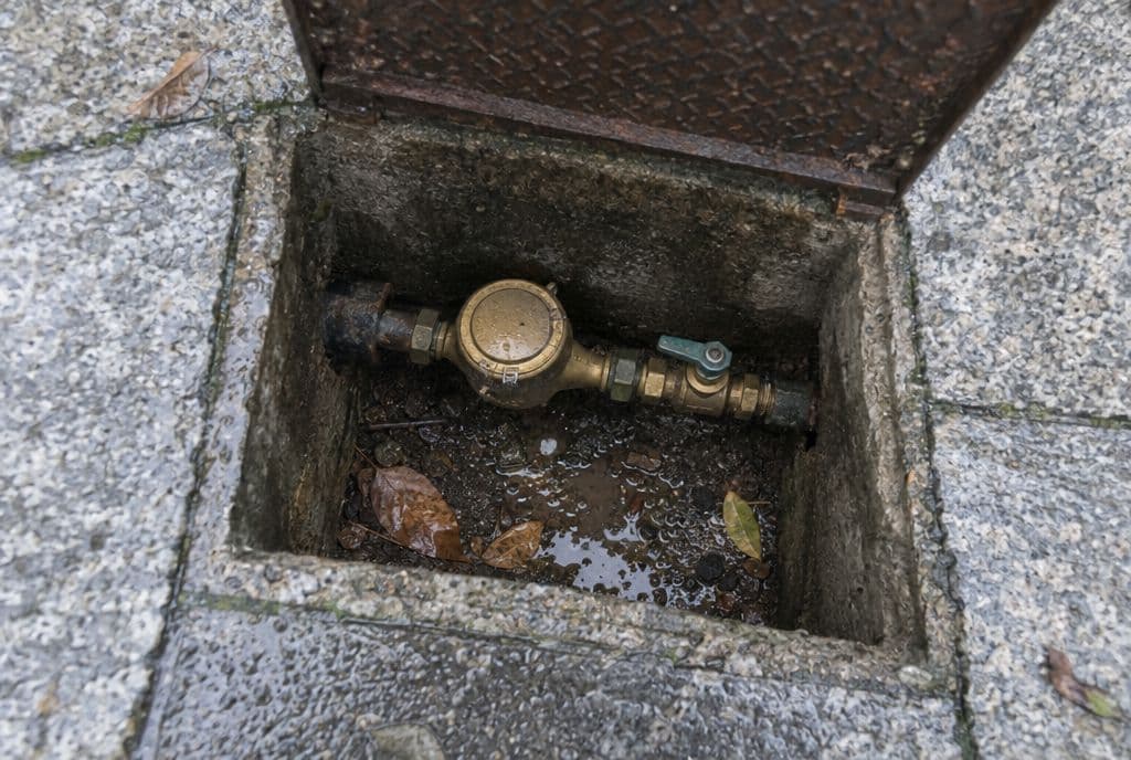 Arqueta de contador de agua abierta en una acera de granito con agua en el fondo